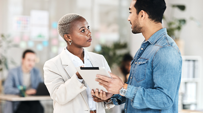 A man and woman in an office discuss information on a mobile tablet.