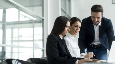 Two businesswomen and a businessman working on a project on a laptop in a well lit conference room.