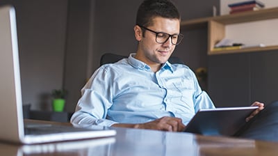 Man in business attire sitting behind a desk in an office, he is looking down at a tablet in his hands.