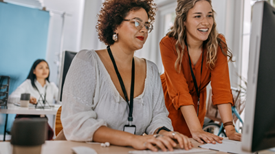 Two women at a desk sharing a computer while having a discussion. Another woman in the background is sitting at desk while working on her computer. 